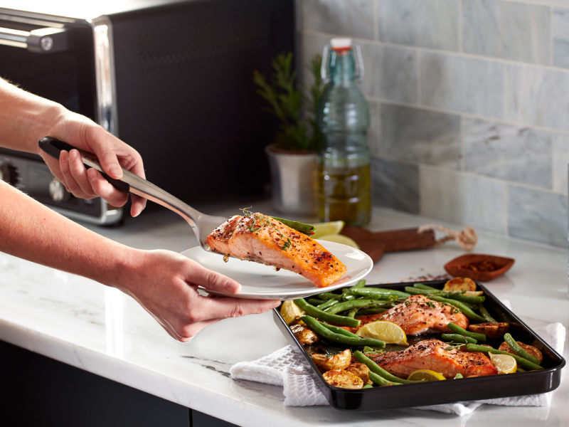 A woman putting freshly baked air fryer salmon on a plate next to a KitchenAid® countertop oven.