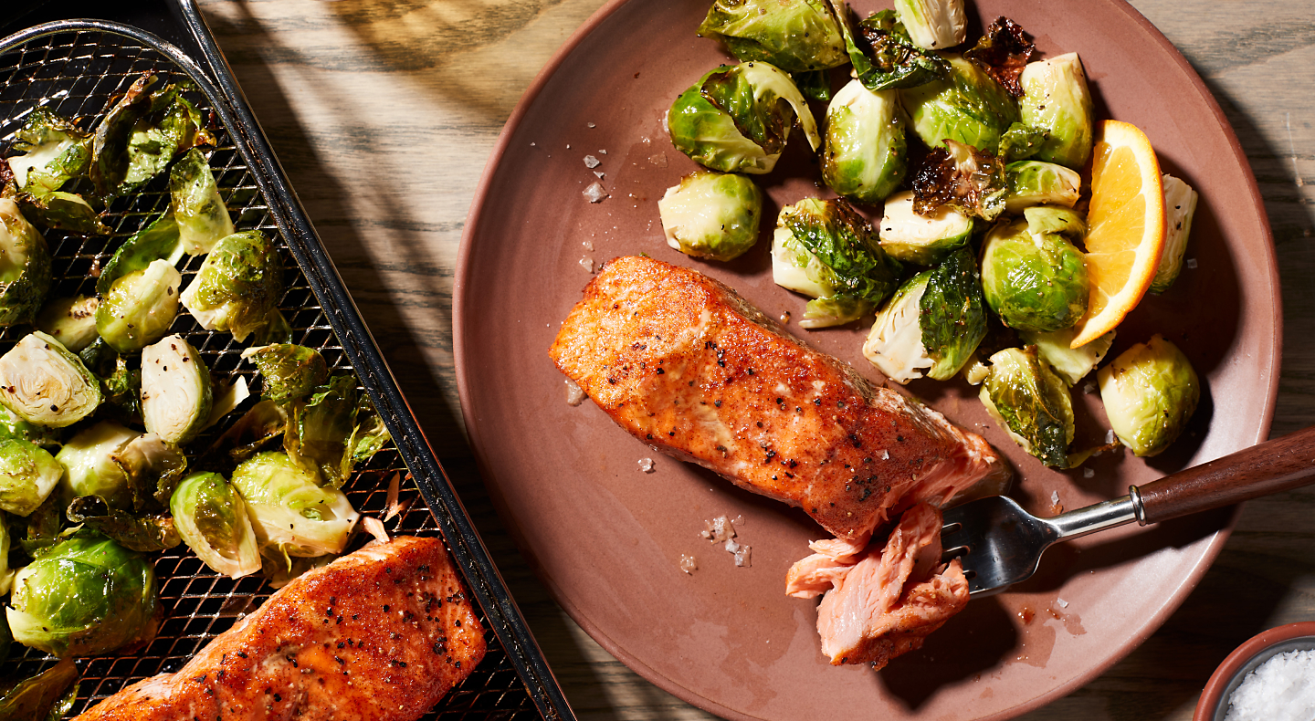 Air fryer salmon and Brussel sprouts on a plate next to an air fry basket with salmon and Brussel sprouts.