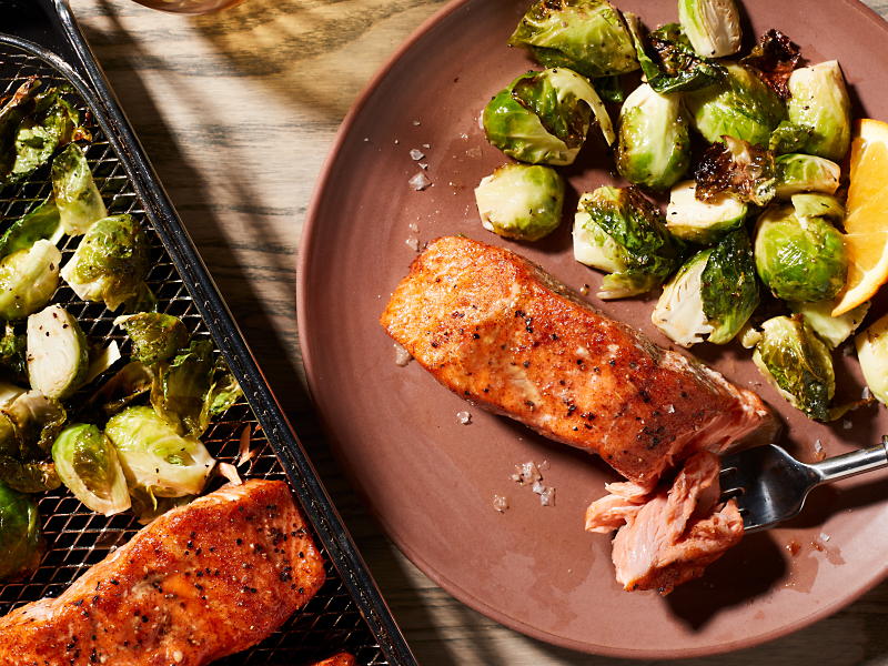 Air fryer salmon and Brussel sprouts on a plate next to an air fry basket with salmon and Brussel sprouts.