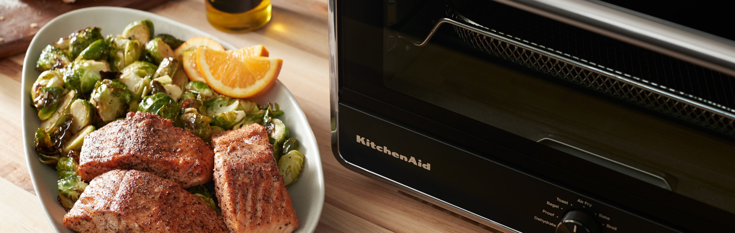 Air fryer salmon in a serving dish with Brussel sprouts next to a KitchenAid® countertop oven with air fry.