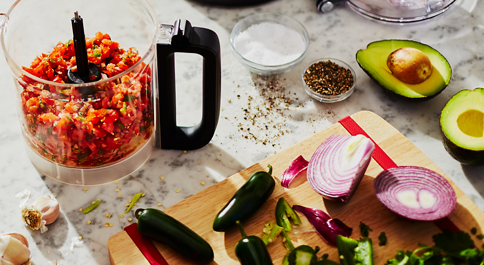 Food processor container with salsa next to cutting board and vegetables 