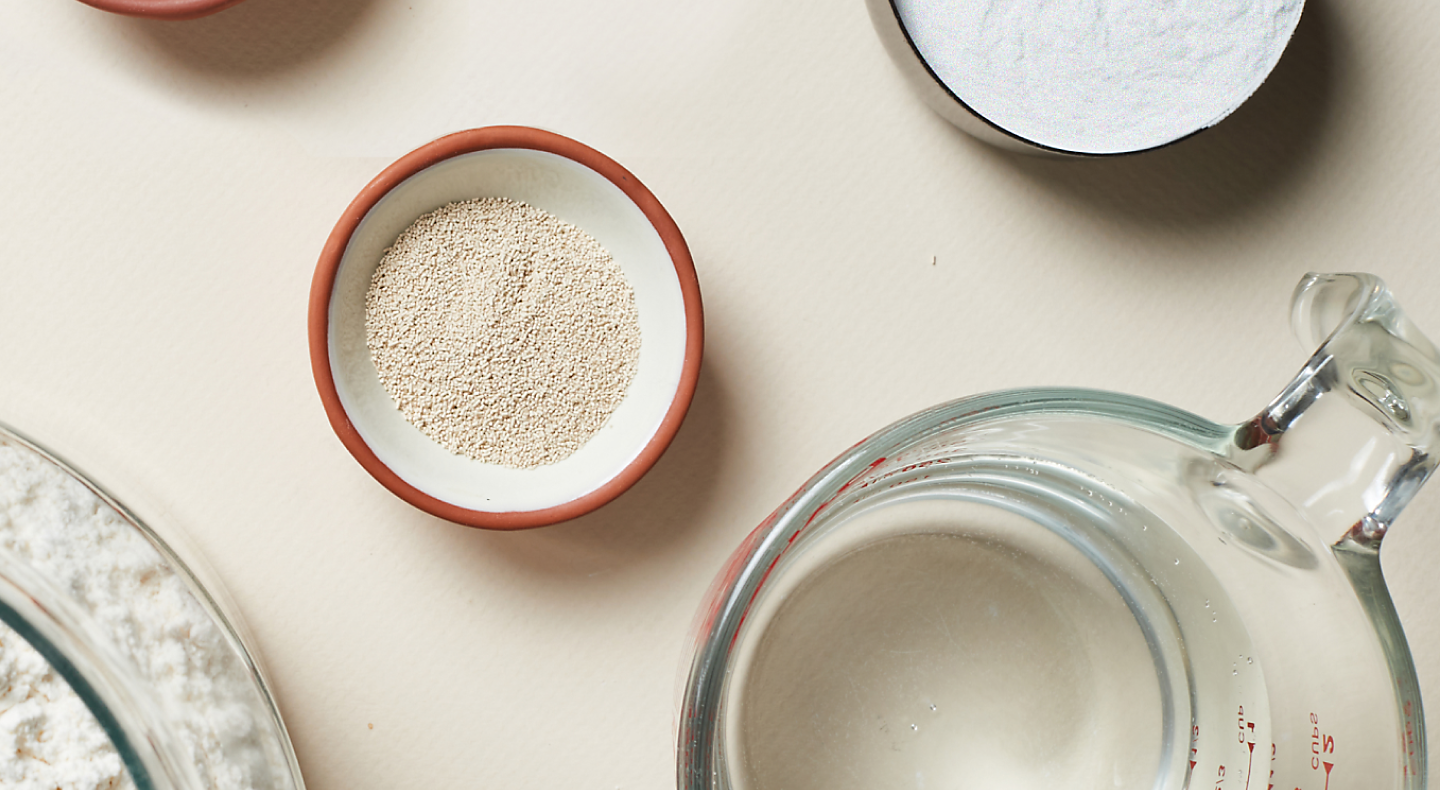 Dry ingredients like yeast and flour in small bowls next to a measuring cup with water