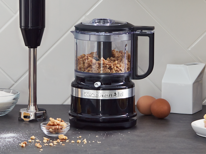 Black KitchenAid® food processor filled with walnuts sitting next to an upright immersion blender on a counter 
