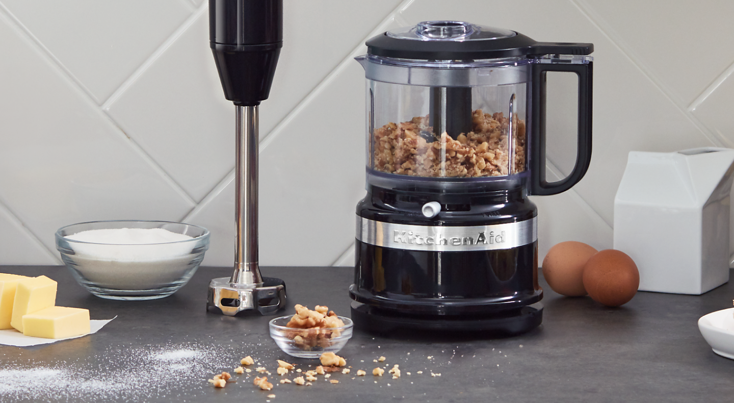 Black KitchenAid® food processor filled with walnuts sitting next to an upright immersion blender on a counter 
