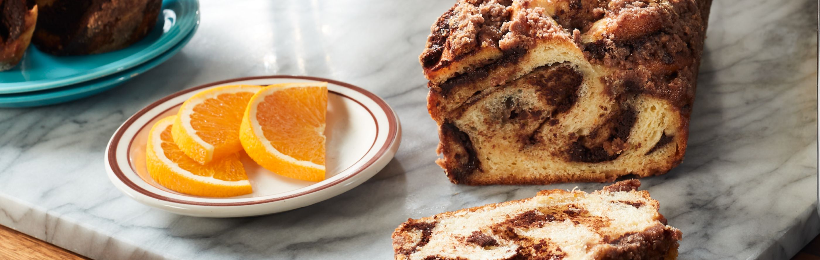 A partially sliced loaf of cinnamon babka on a marble cutting board next to a plate of orange slices 