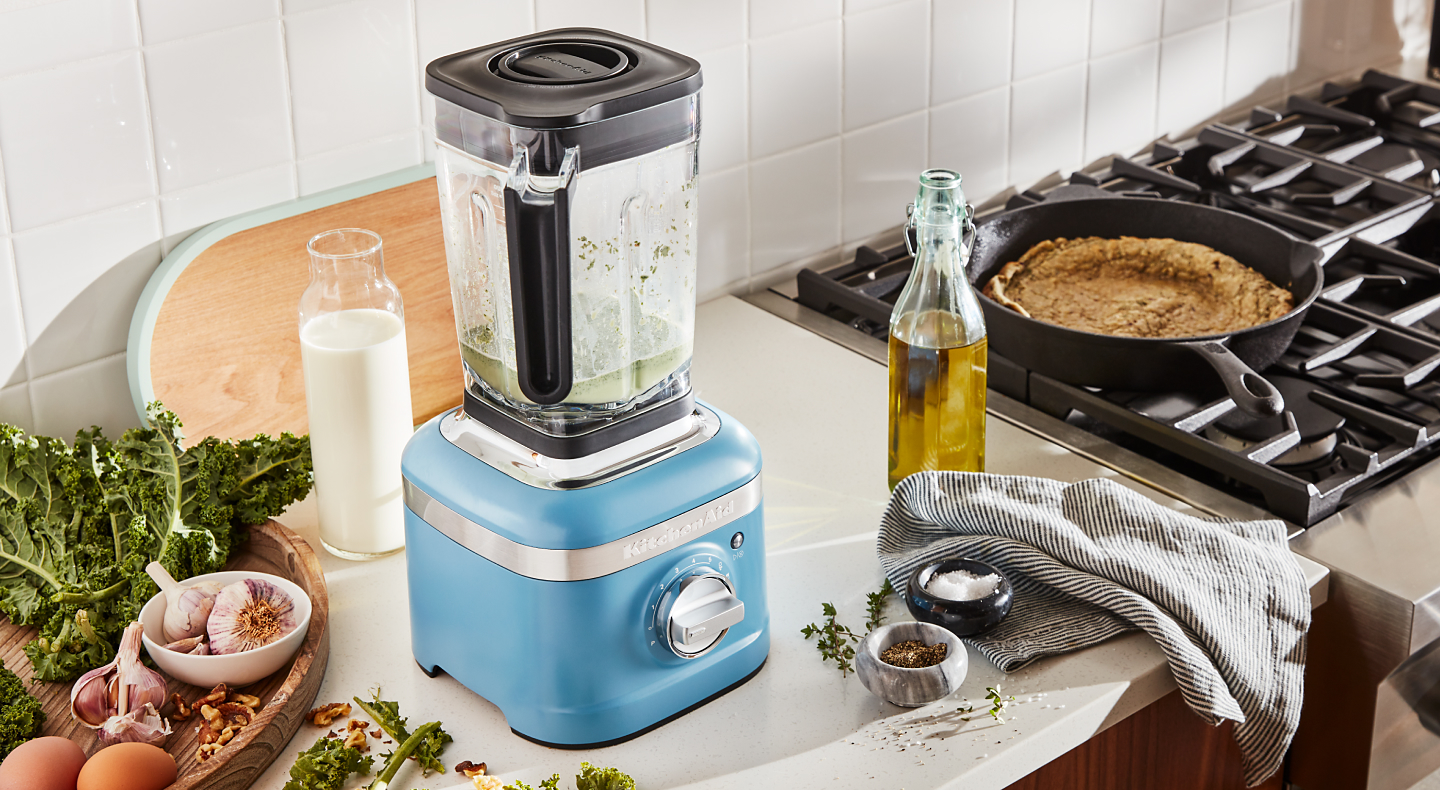 A KitchenAid® blender surrounded by various herbs and vegetable oil.