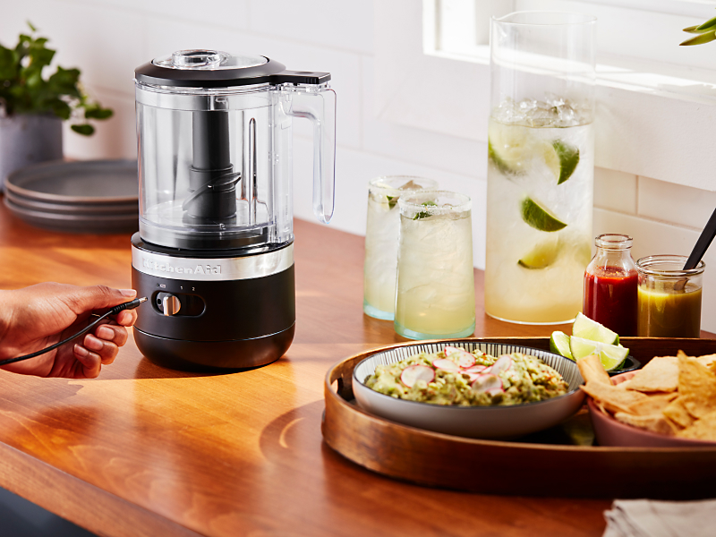 A cordless KitchenAid® food chopper next to a platter of guacamole and tortilla chips and a pitcher and glasses of margaritas. 