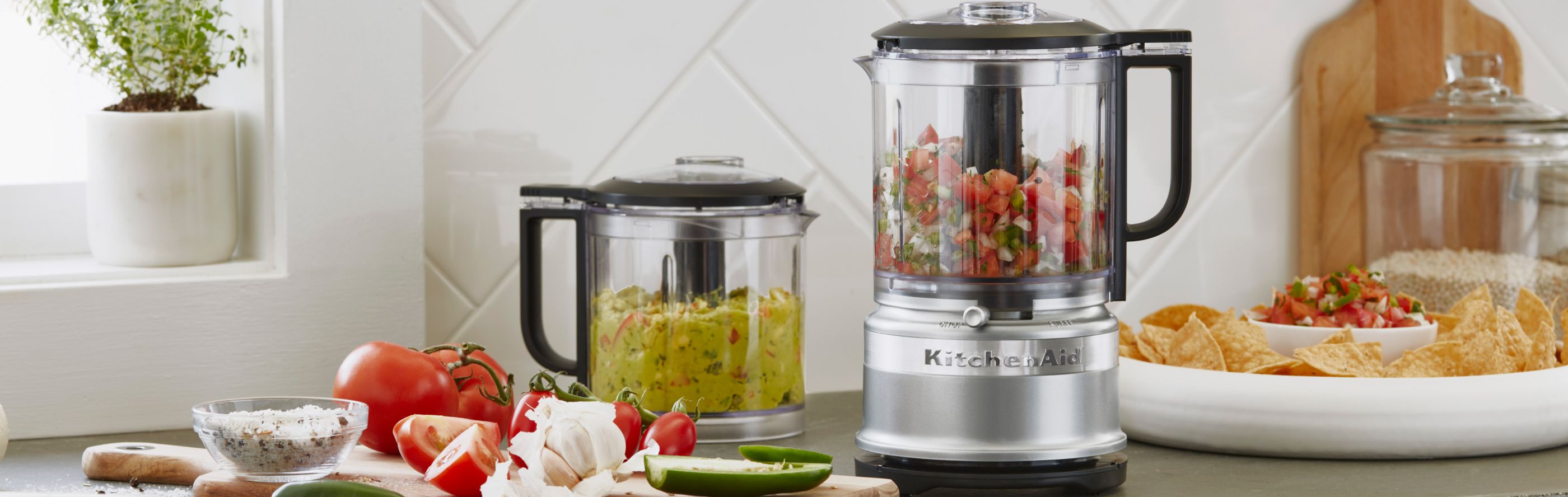 A KitchenAid® food processor and food chopper with freshly made salsa and guacamole on a modern kitchen counter.
