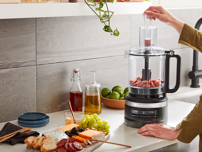 Person dropping food into the feed tub of a black KitchenAid® food processor