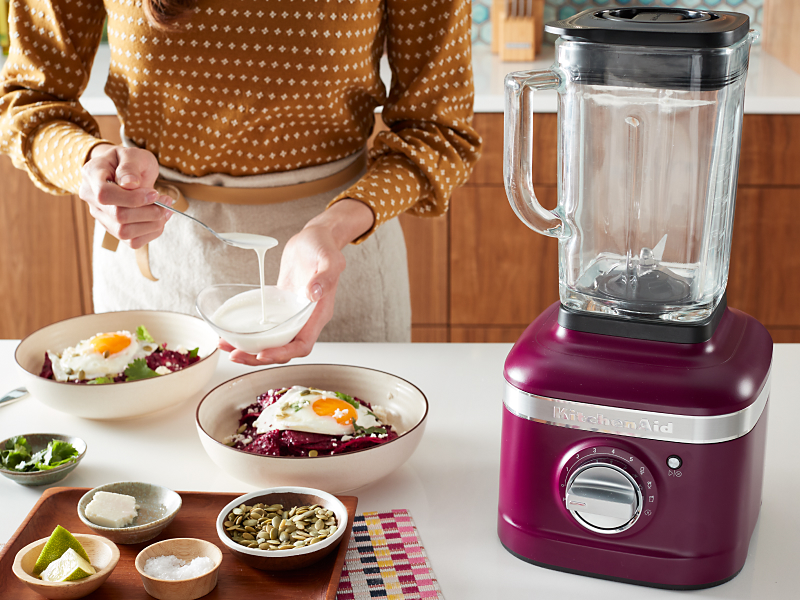Person topping a harvest bowl with sauce next to a KitchenAid® blender