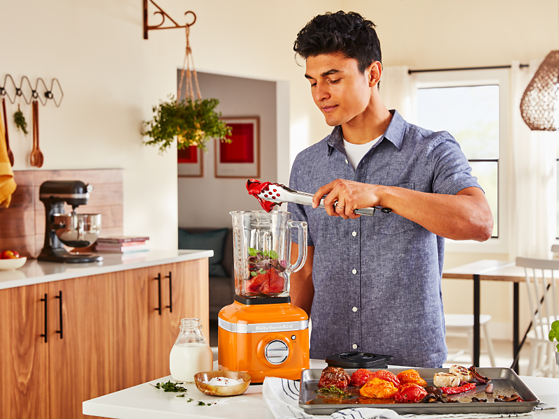 Person adding roasted vegetables into an orange KitchenAid® blender