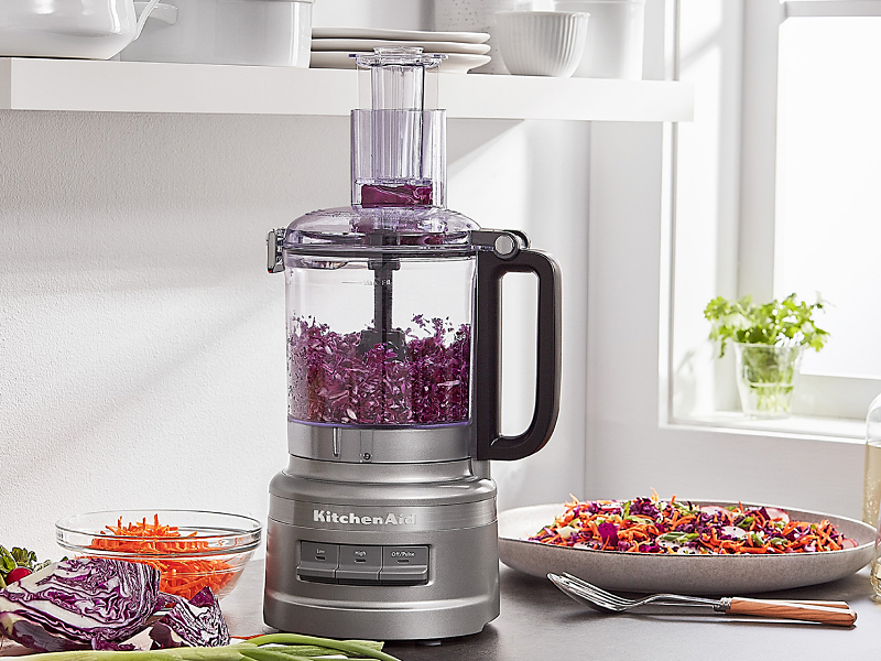 Shredded purple cabbage in a KitchenAid® food processor on a kitchen counter next to a plate of food