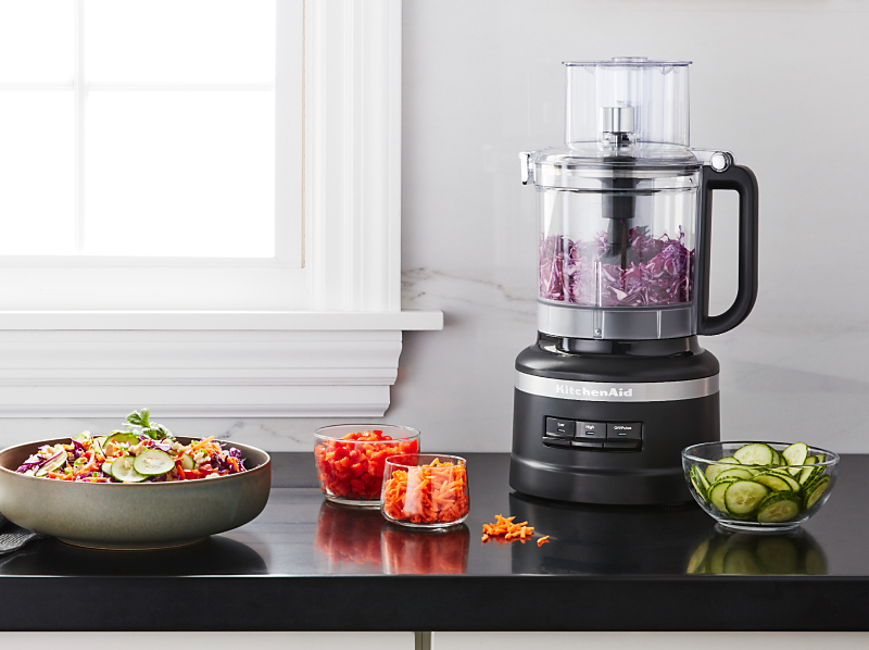 Shredded purple cabbage in a KitchenAid® food processor on a kitchen counter next to other shredded vegetables