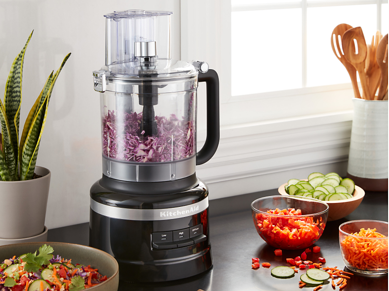Shredded purple cabbage in a KitchenAid® food processor on a kitchen counter next to other shredded vegetables