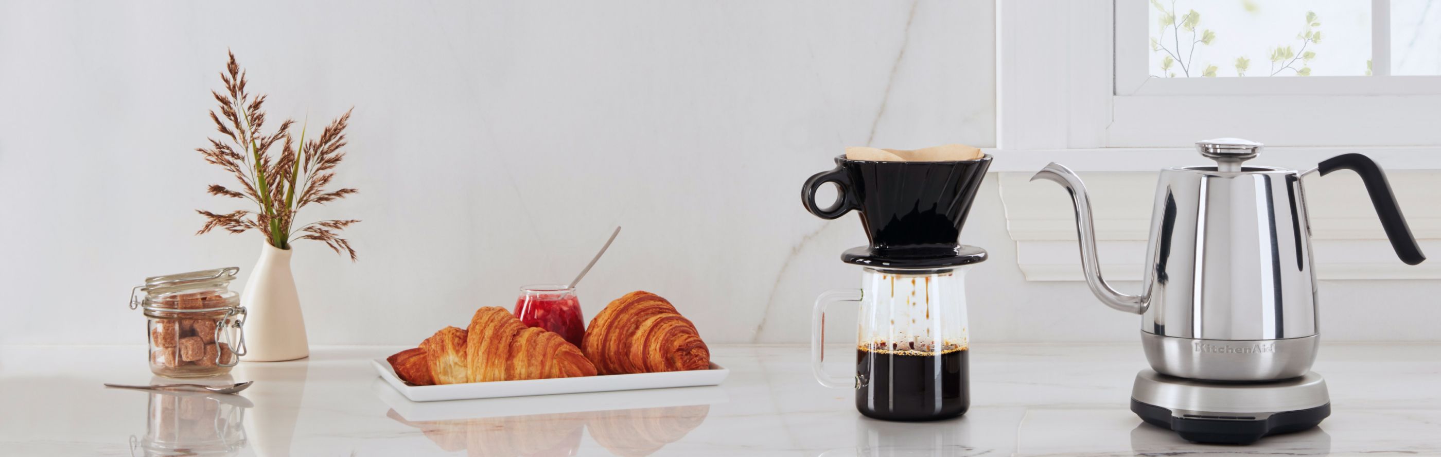 Silver KitchenAid® electric kettle on countertop with coffee and plate of croissants