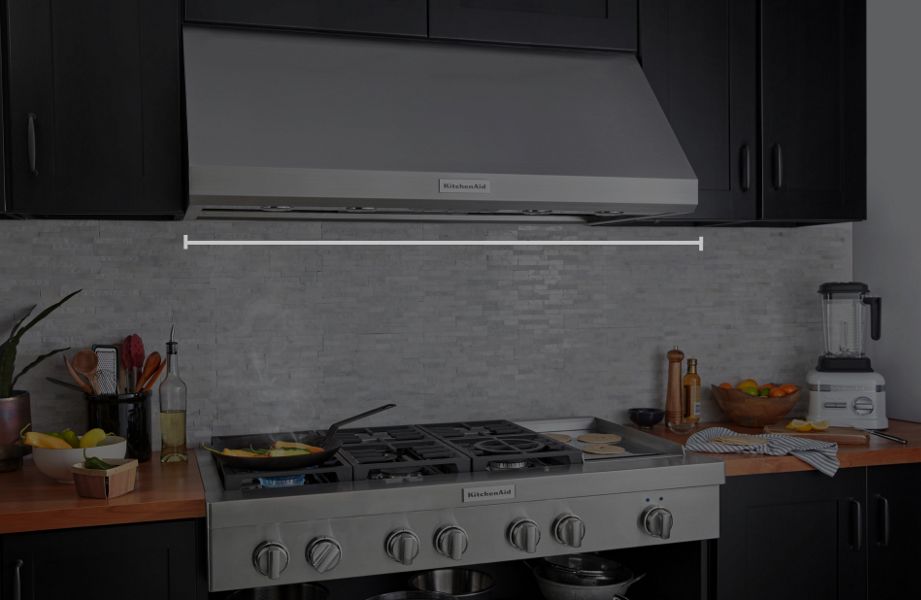 Under-cabinet KitchenAid® hood above a KitchenAid® gas range with food cooking in pan and an animated hood width measurement overlay