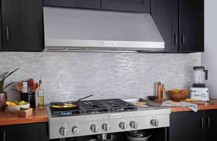 Under-cabinet KitchenAid® hood above a KitchenAid® gas range with food cooking in pan and an animated hood width measurement overlay