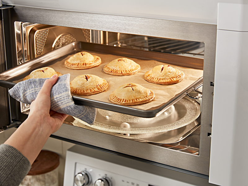A person removing a sheet pan of baked pastries from a KitchenAid® microwave.