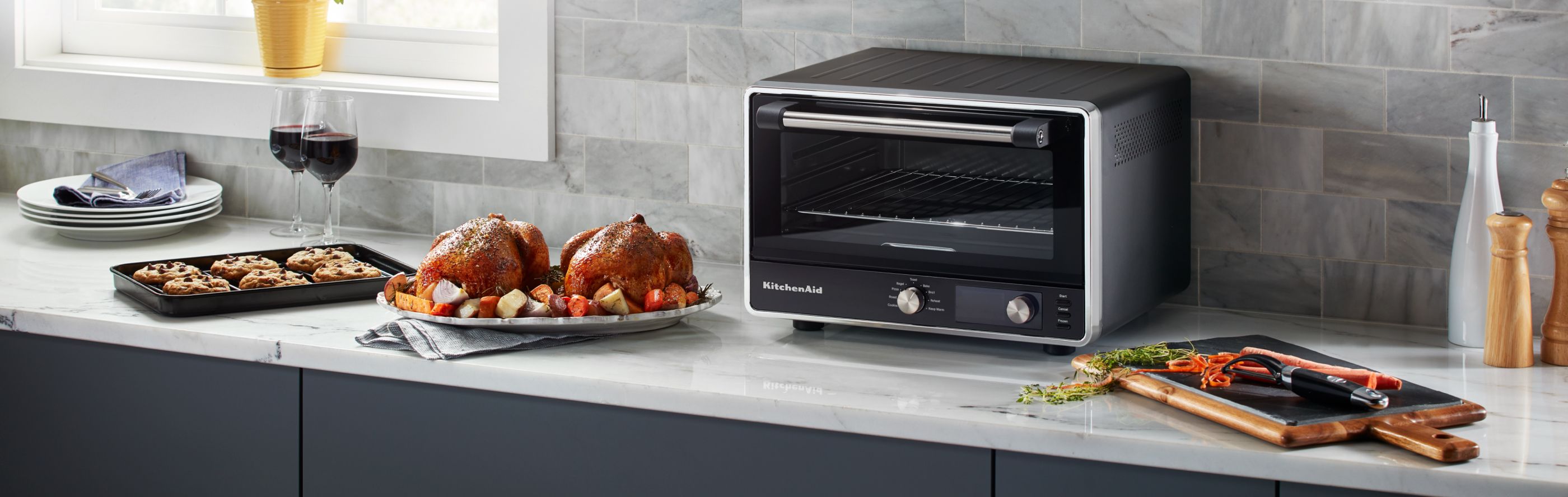 KitchenAid® countertop oven next to roasted chicken and a tray of cookies