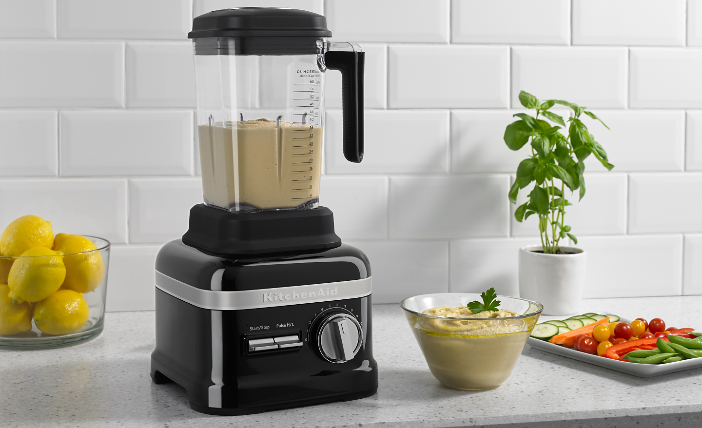 A KitchenAid® blender with hummus on a modern kitchen counter next to fresh vegetables.