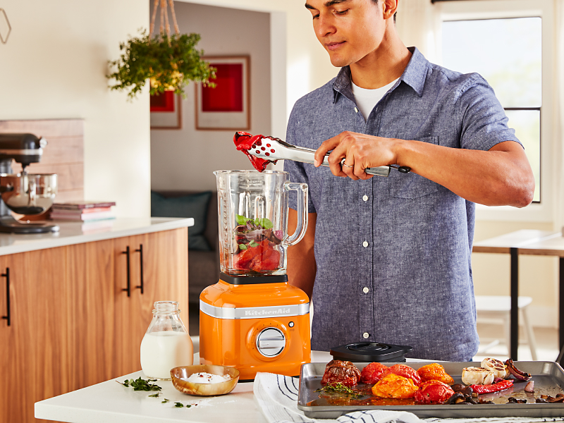A man putting roasted vegetables into a KitchenAid® blender in a modern kitchen.