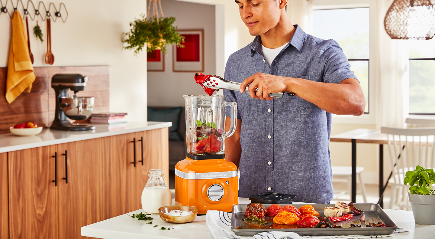 A man putting roasted vegetables into a KitchenAid® blender in a modern kitchen.