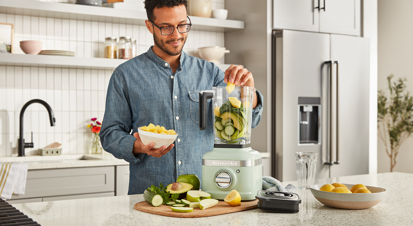 A man adding vegetables to a KitchenAid® blender in a modern kitchen.