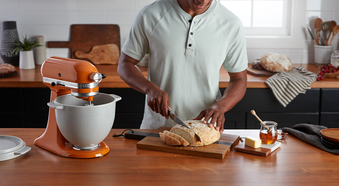 Person slicing crispy bread on cutting board next to orange KitchenAid® stand mixer