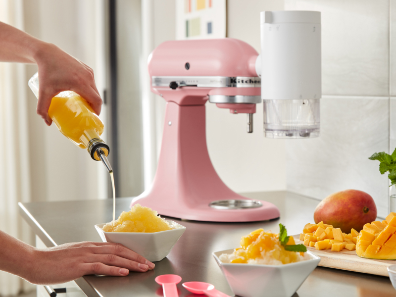 Person pouring syrup over shaved ice next to a KitchenAid® Shave Ice Attachment on a pink KitchenAid® stand mixer 