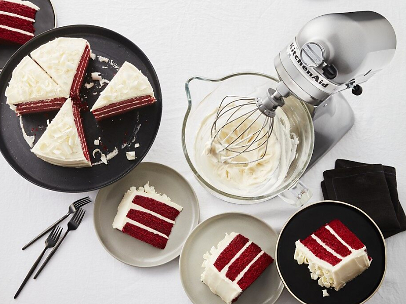 A KitchenAid® stand mixer with whipped white frosting next to a sliced red velvet cake.