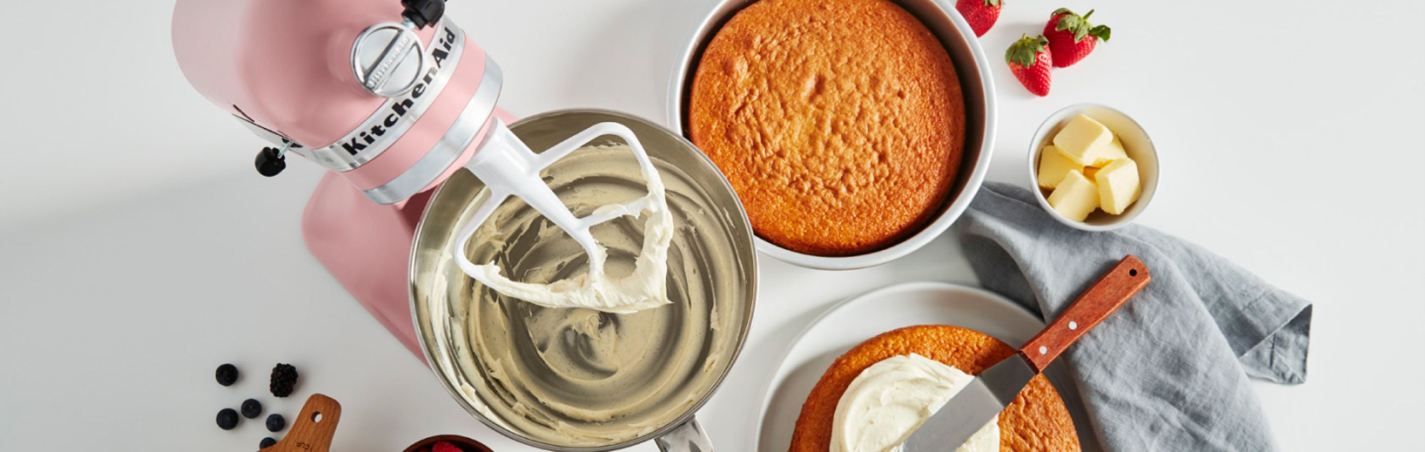 A KitchenAid® stand mixer with cake frosting next to a couple of baked cakes ready for frosting. 