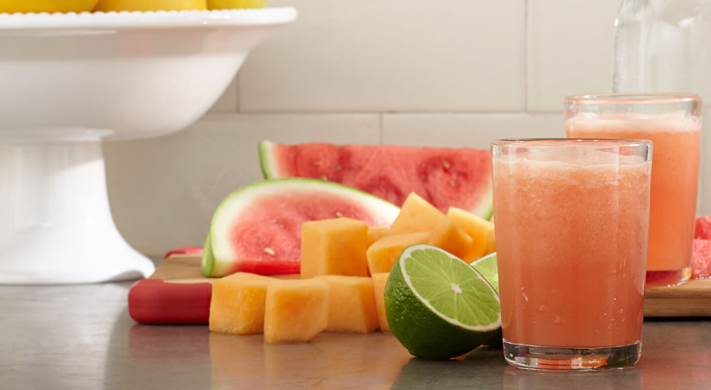 A frozen mocktail next to limes, watermelon slices and cantaloupe chunks.