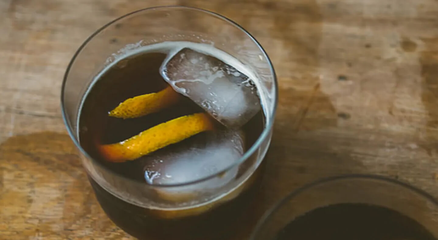 Two ice cubes in a glass of soda with an orange peel garnish.