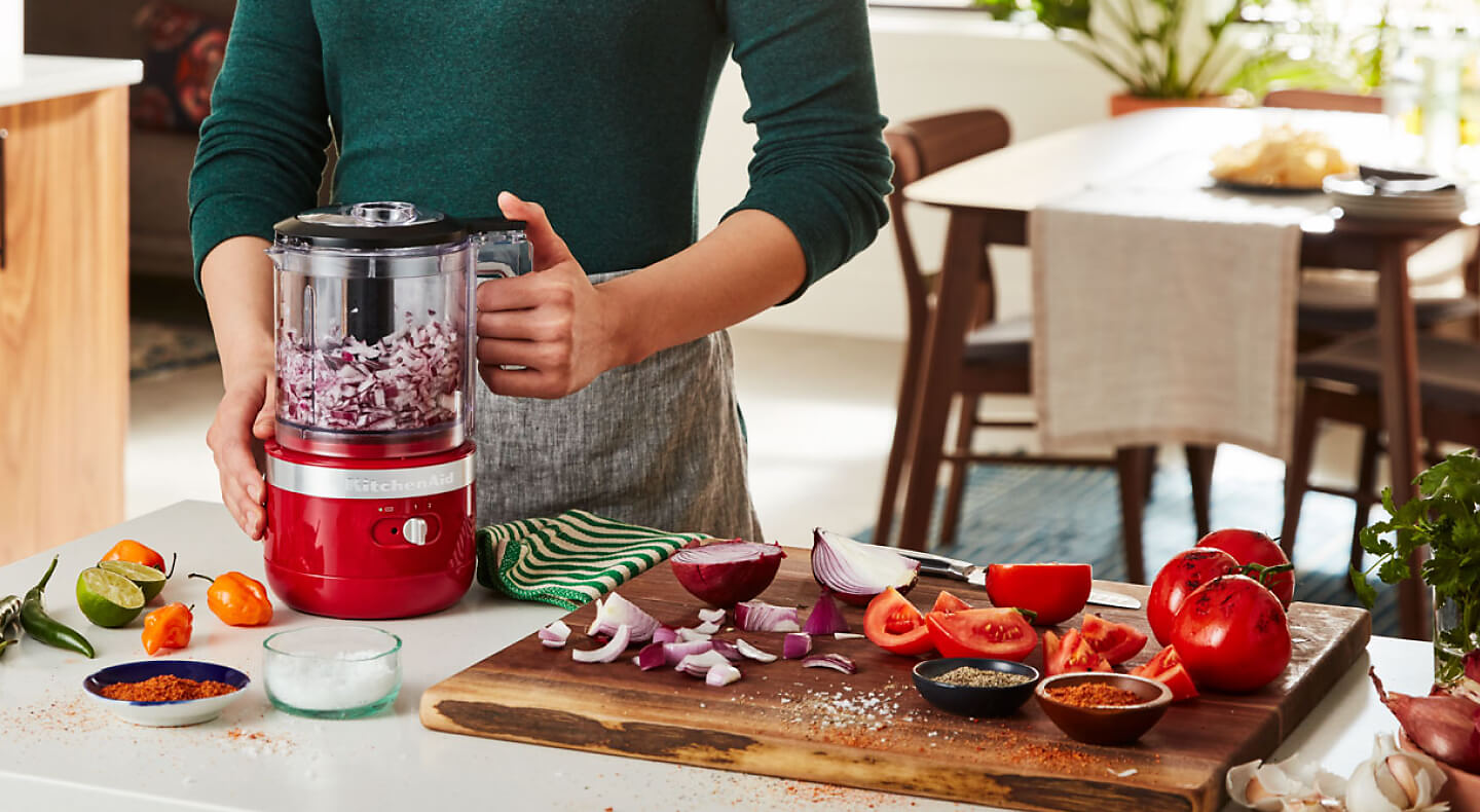 A person chopping red onions in a KitchenAid® food chopper next to a cutting board with red onions, tomatoes and spices