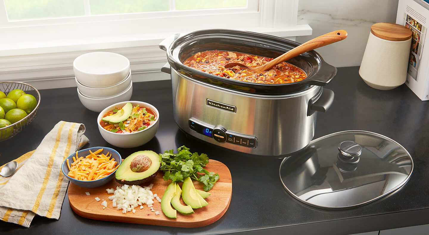 A KitchenAid® slow cooker with chicken chili, next to a cutting board with cheese, avocado, cilantro and onions 