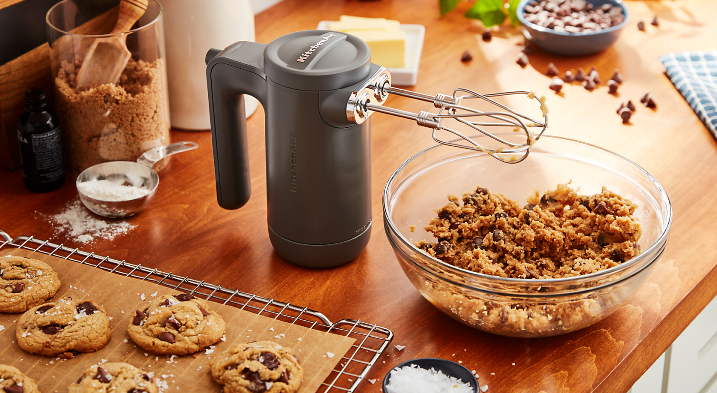A cordless hand mixer on a countertop with freshly baked cookies and a bowl of cookie batter