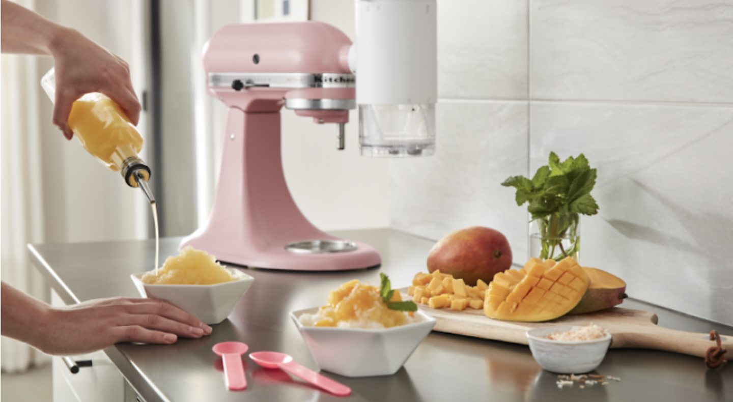 Person pouring syrup on a bowl of shaved ice surrounded by KitchenAid® stand mixer and chopped mangoes
