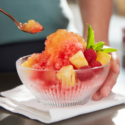 Person placing spoon in shave ice dessert topped with chopped fruit and syrup