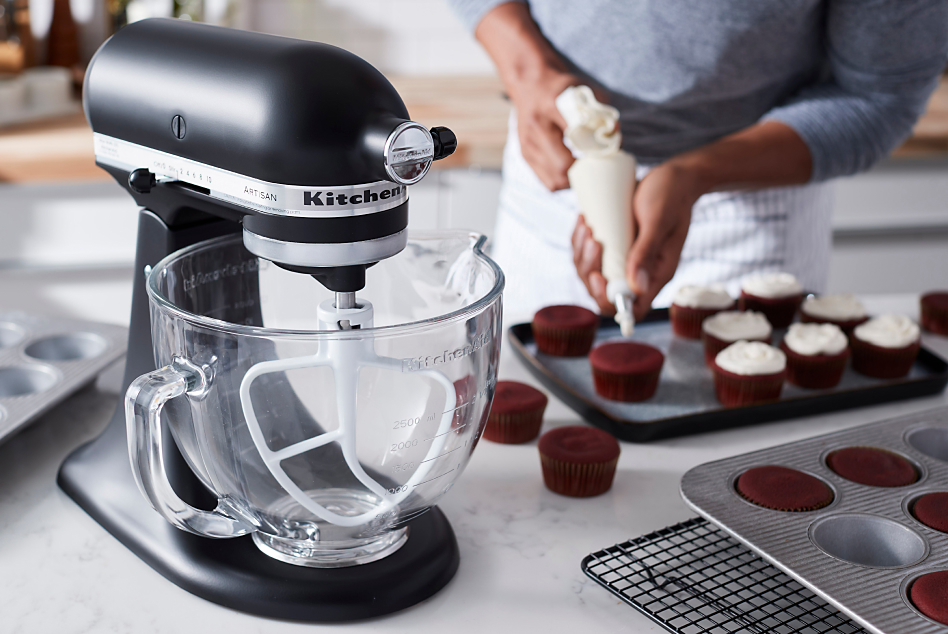 Black stand mixer with glass bowl and man frosting cupcakes in background