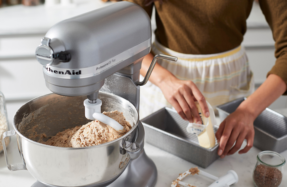 Silver stand mixer kneading tough dough and woman greasing bread pan in background