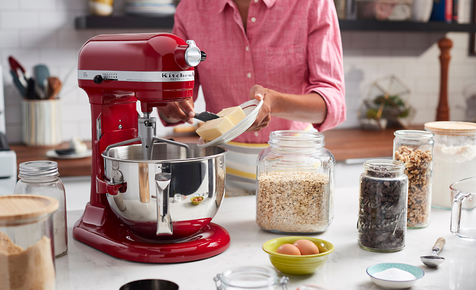 Red bowl-lift stand mixer on counter with jars of ingredients, woman adding butter to bowl
