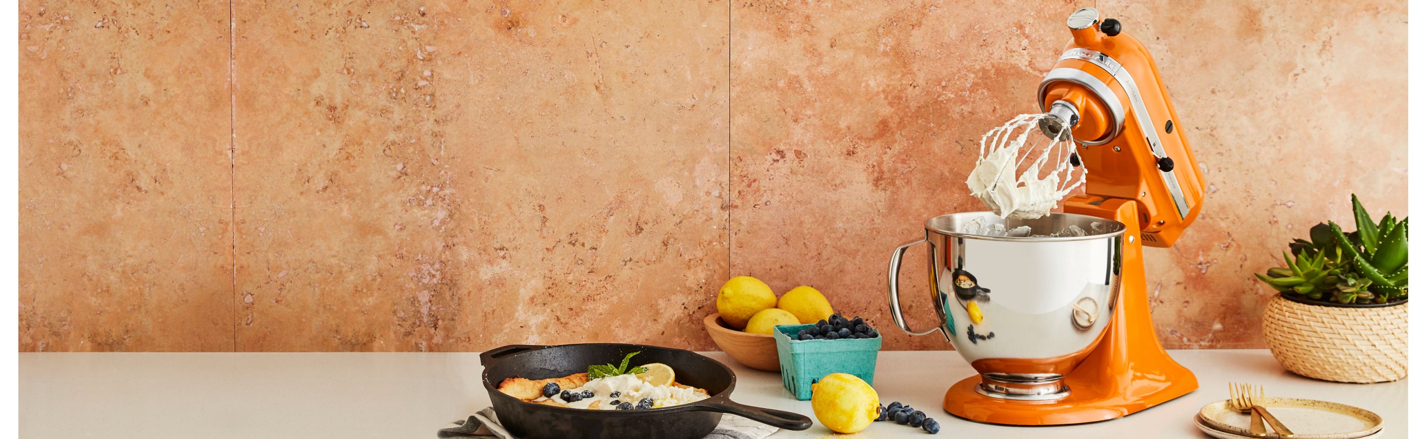 An orange KitchenAid® stand mixer, fruit and a cast iron pan.