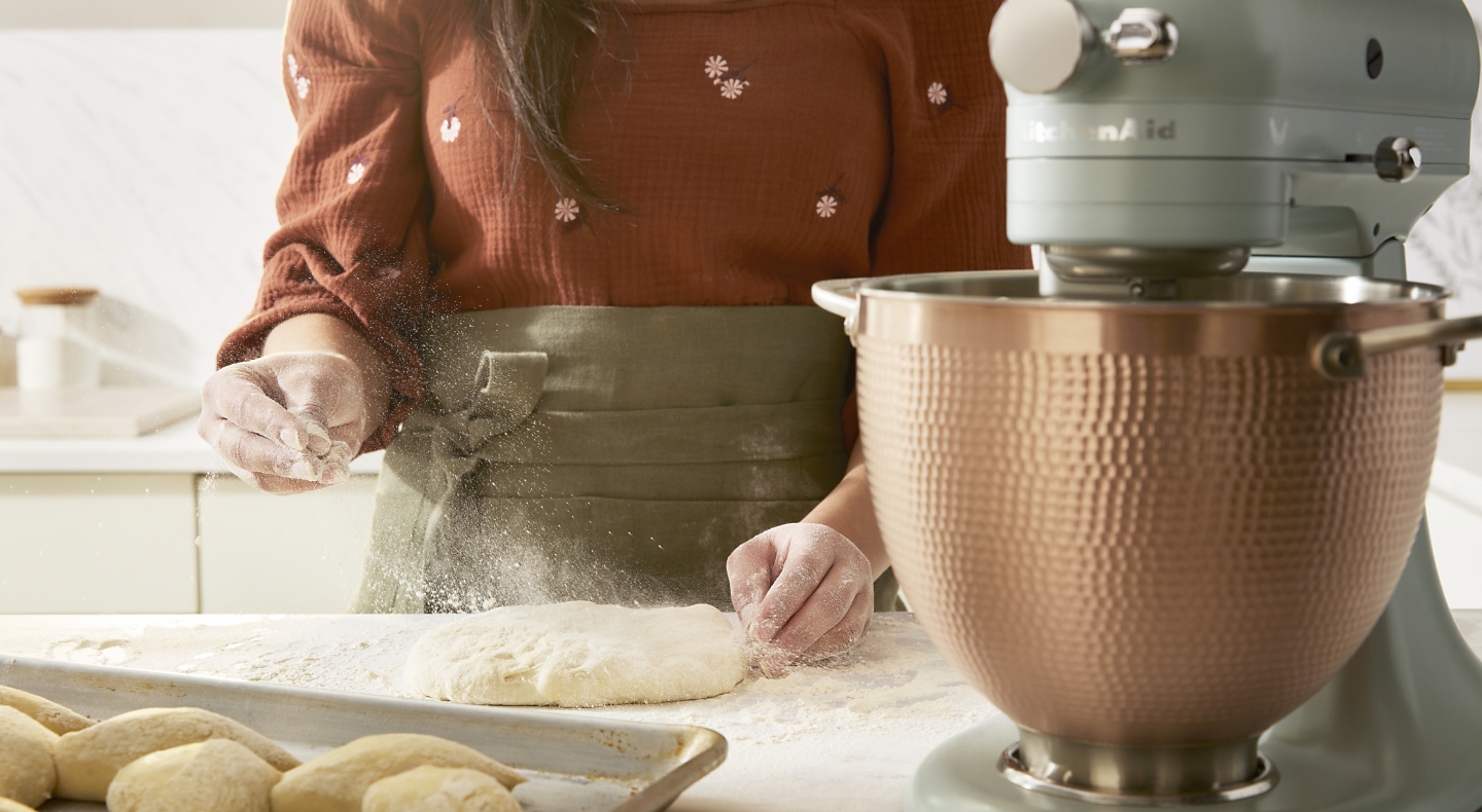 A person sprinkling dough with flour next to a KitchenAid® stand mixer.