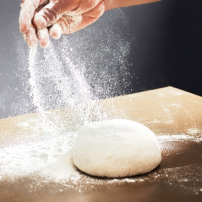 A person sprinkling flour onto a smooth kneaded dough ball on a wooden countertop