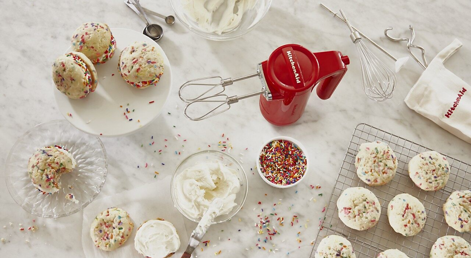 KitchenAid® hand mixer on countertop with frosting and sprinkles for homemade cupcakes.