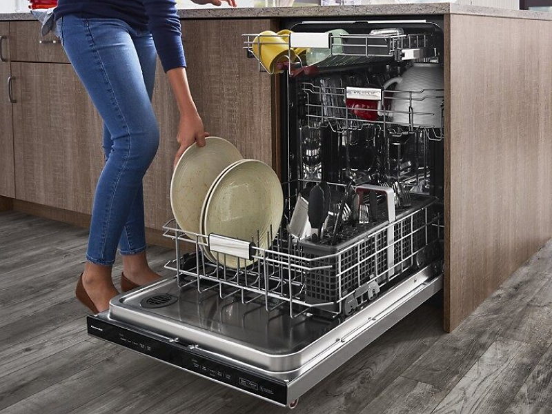 A closeup of a person loading plates onto the bottom rack of a stainless steel KitchenAid® dishwasher.