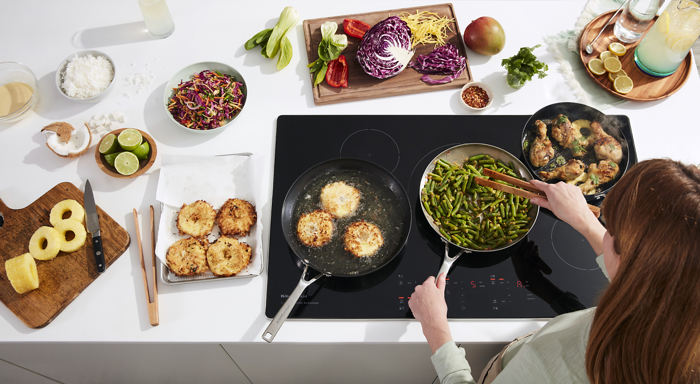 Person sauteeing green beans on electric cooktop surrounded by ingredients