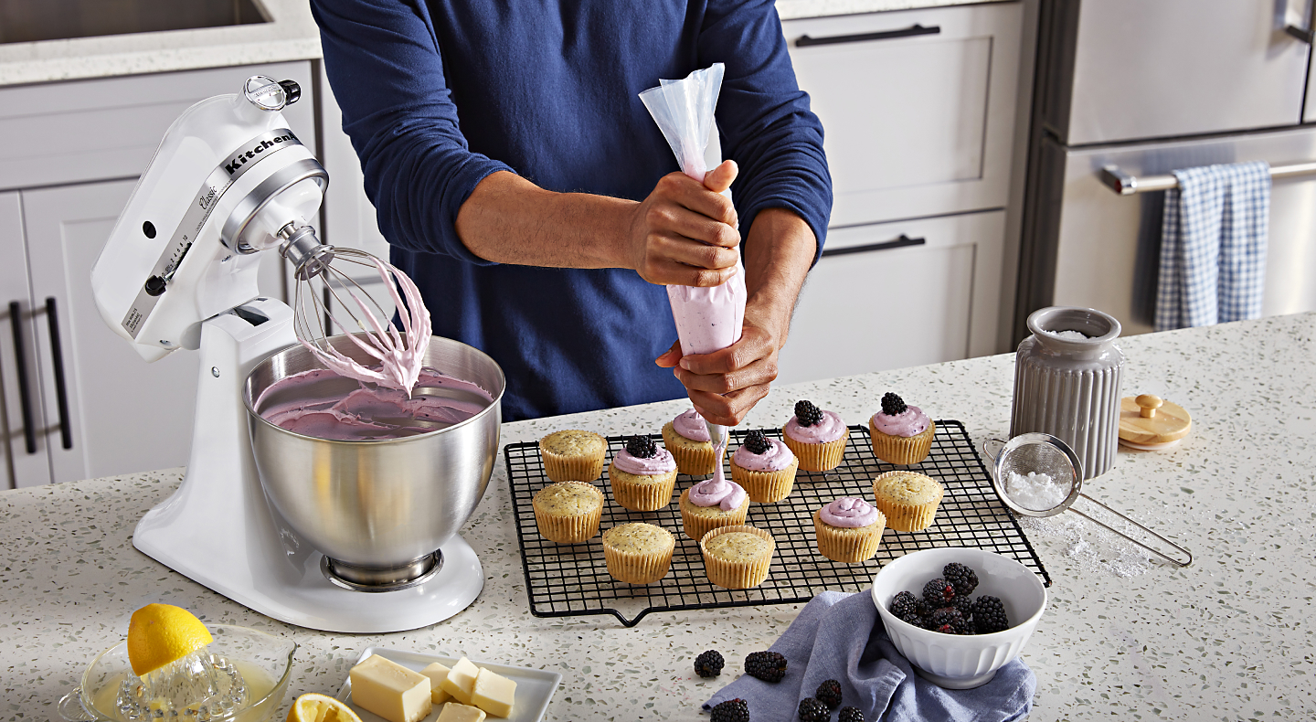 Person piping frosting onto cupcakes next to white stand mixer full of frosting