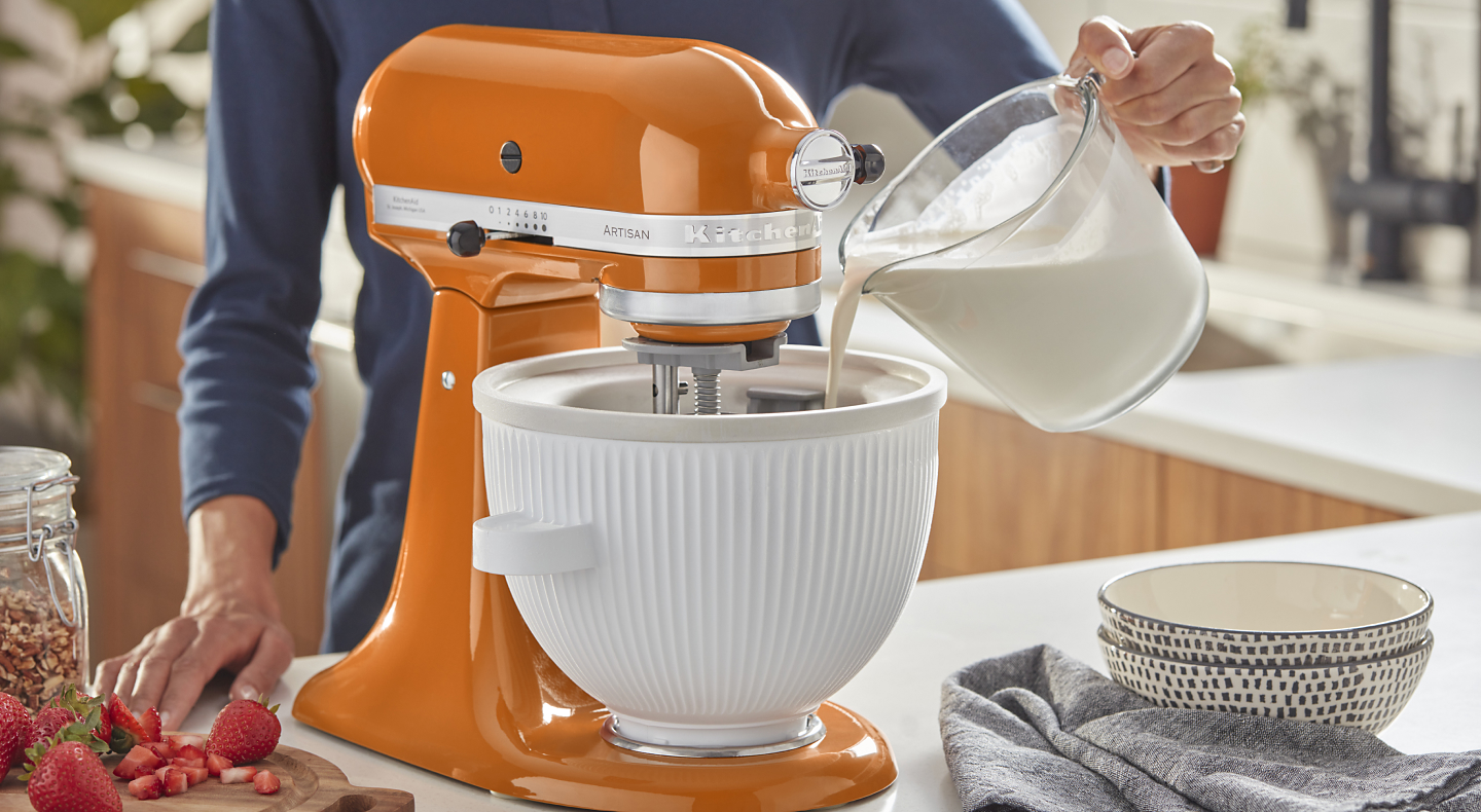 Person pouring milk into the white bowl of an orange stand mixer