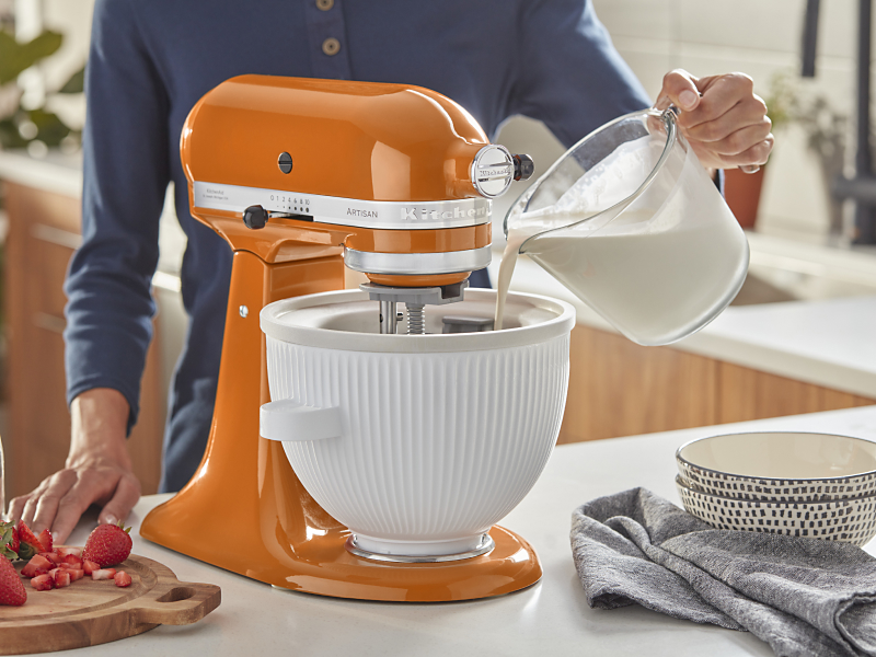 Person pouring milk into the white bowl of an orange stand mixer